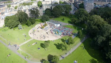 Aerial-shot-of-the-children's-play-park-in-the-meadows-in-Edinburgh,-filled-with-children-playing,-on-a-sunny-Summer-day-|-Edinburgh,-Scotland-|-4k-at-30-fps