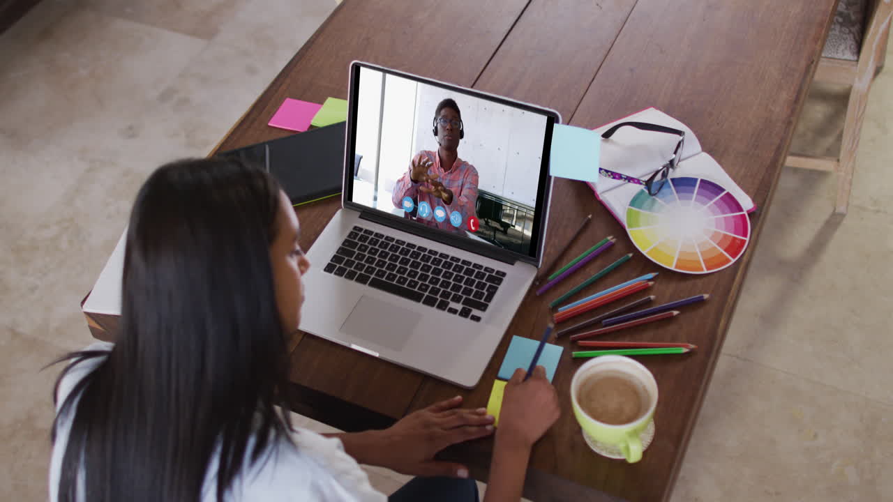 Mujer caucásica usando una laptop en videollamada con un colega, tomando notas