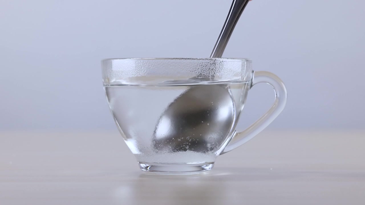Un cucchiaio di metallo mescola polvere bianca nell'acqua calda all'interno di una tazza di vetro trasparente