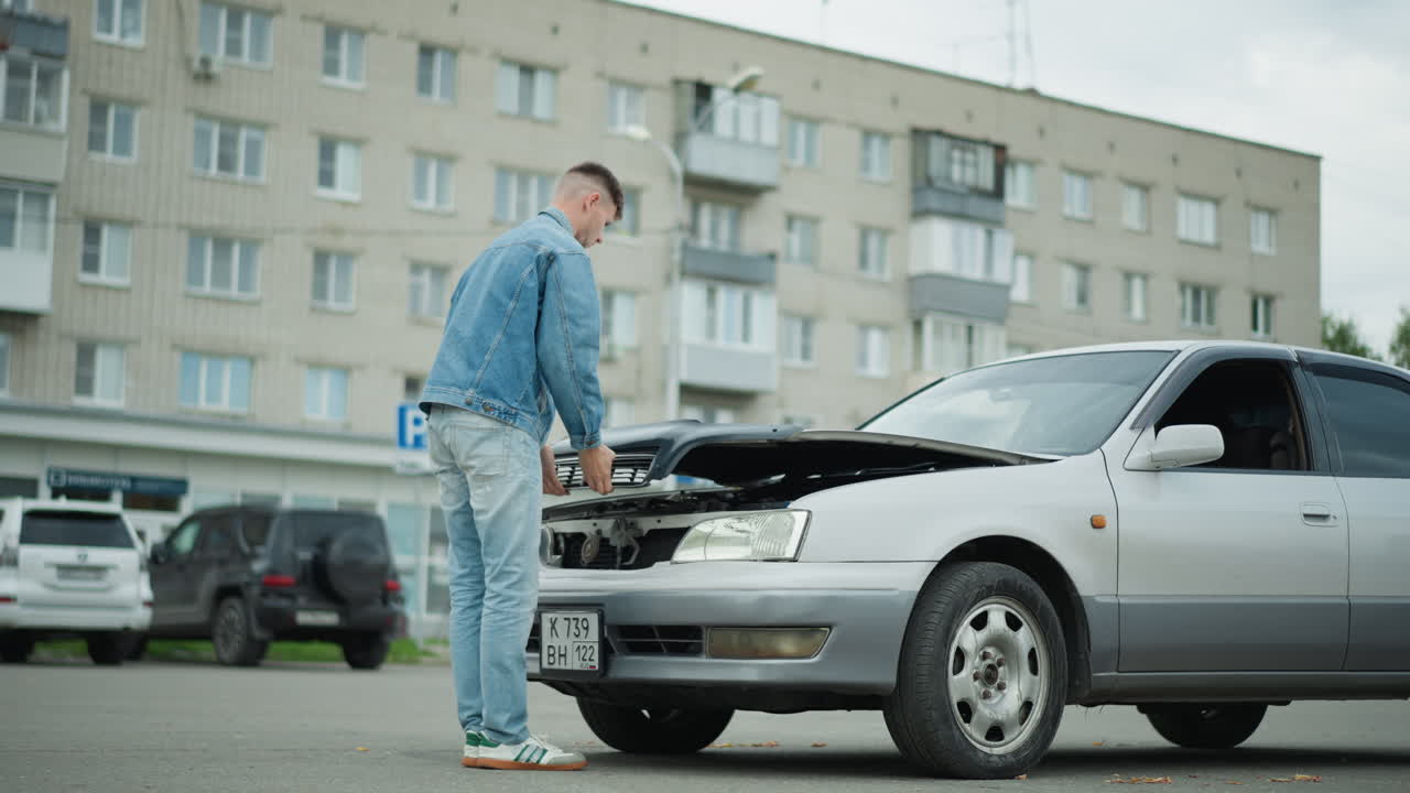Un hombre blanco joven examina el compartimento del motor en un estacionamiento residencial, con el capó levantado y las manos explorando los componentes. Viste un conjunto de tela vaquera, tiene una expresión contemplativa y está junto a un sedán plateado con daños leves en la parte delantera.