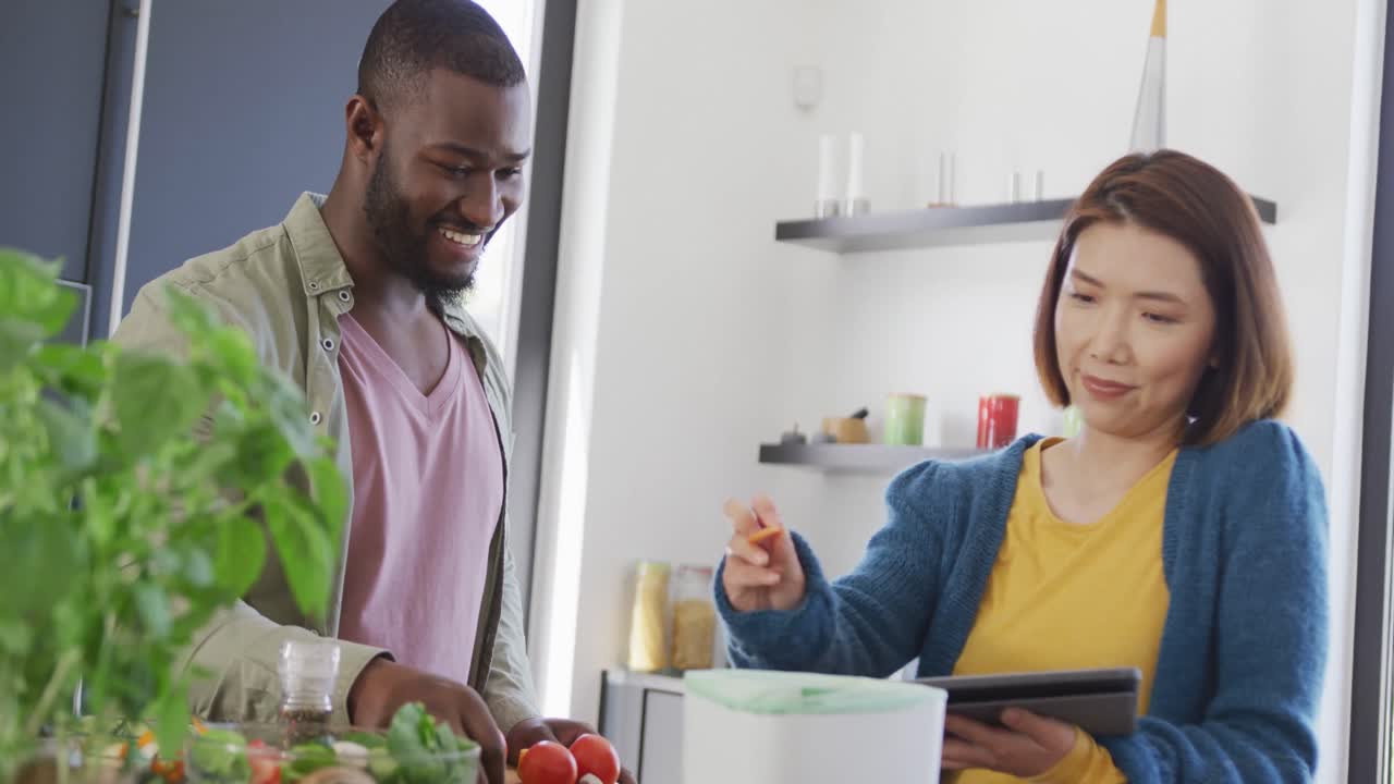 Video of happy diverse couple preparing food, using tablet and composting vegetables in kitchen