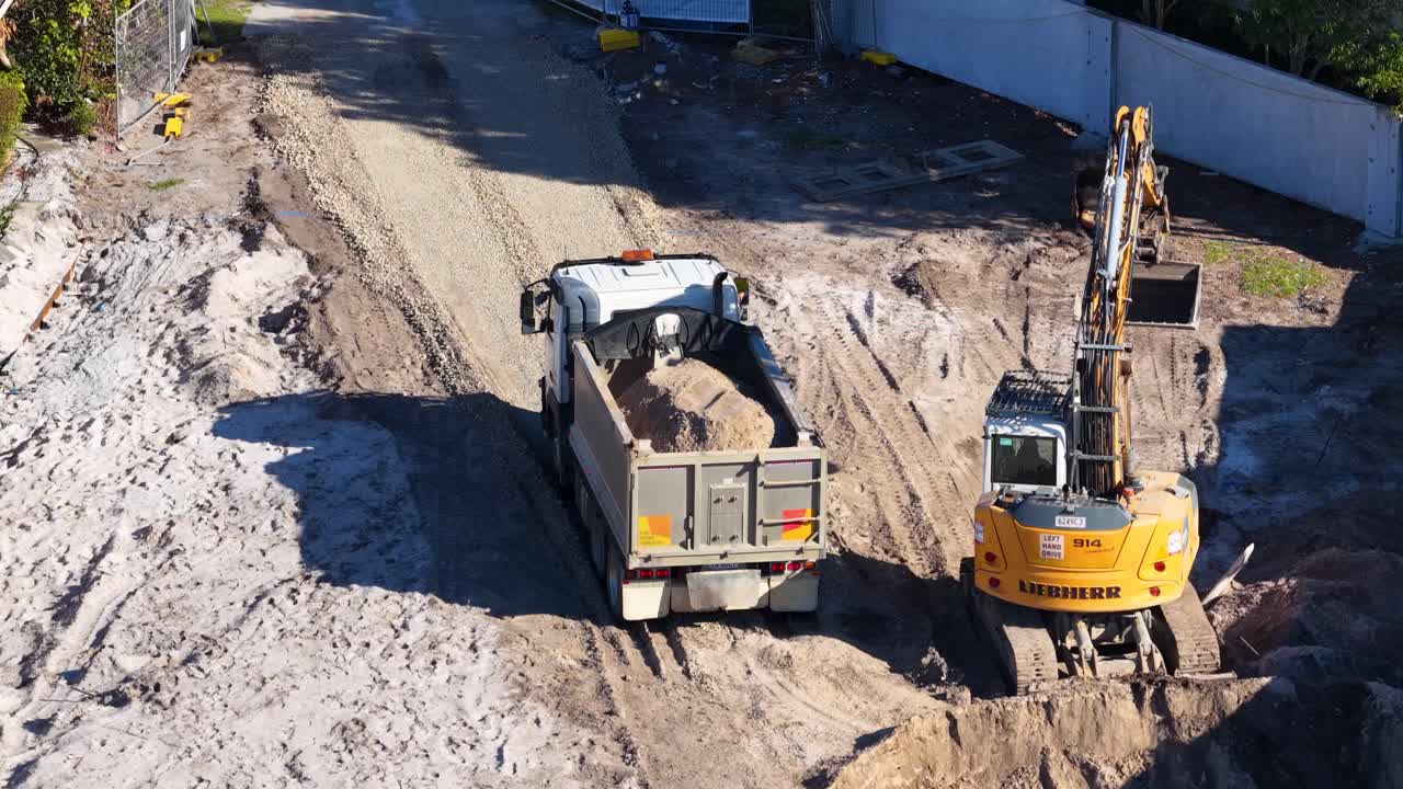 Excavator loads soil into dump truck, bright daylight, aerial view, construction site, steady camera