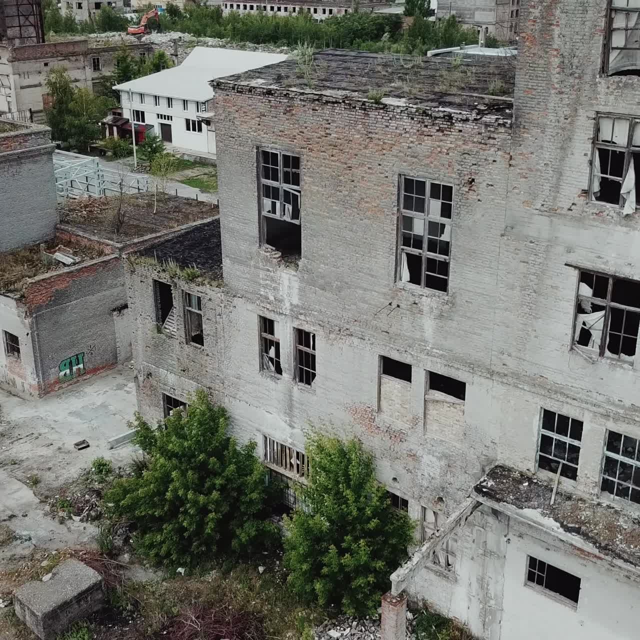 Aerial view. Ruined old building, tilt down to the dry grass and bushes. Abandoned factory halls with broken glass on windows. Apocalyptic scene with destroyed warehouse.