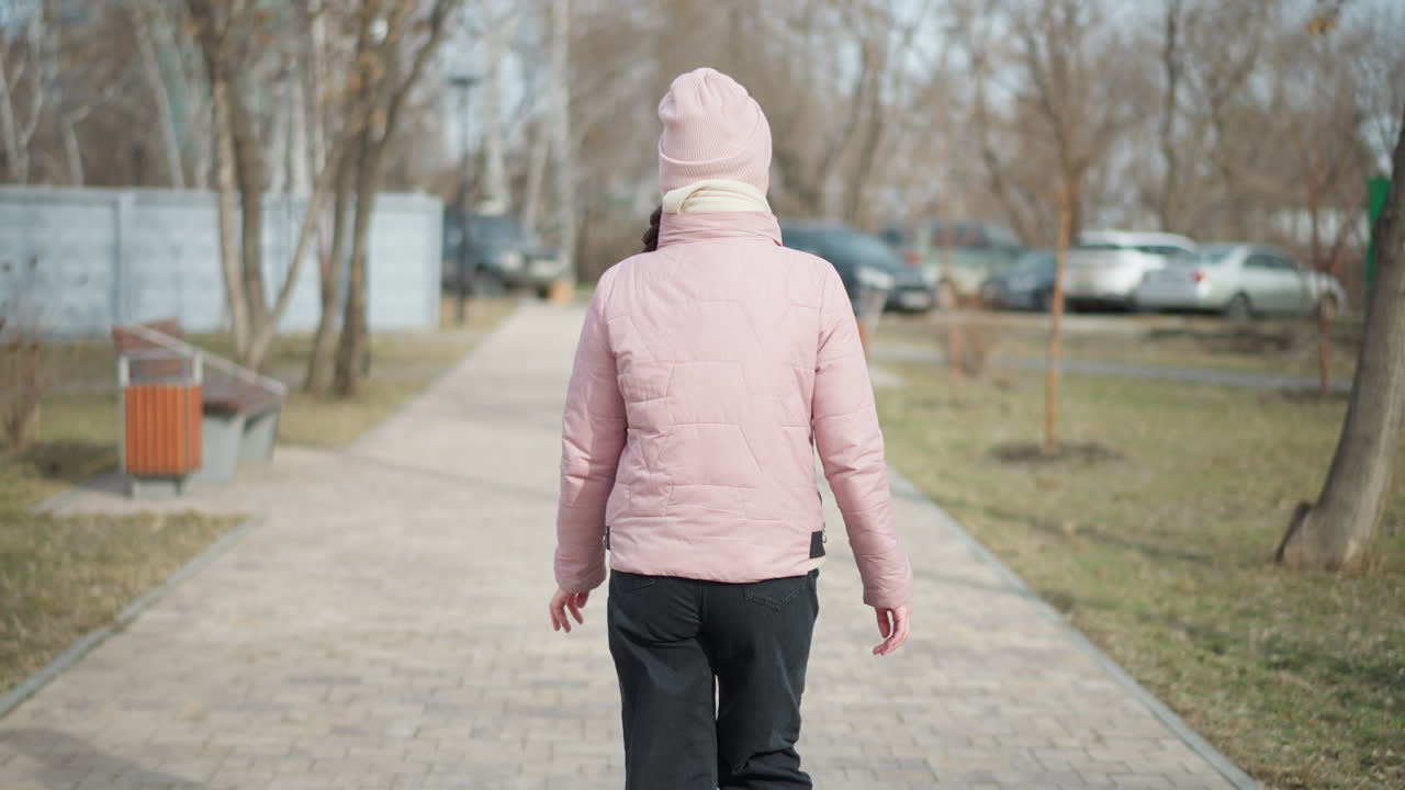 Woman dressed in pink winter jacket and hat walks away on paved path in quiet park during cold day, hands relaxed, surrounded by leafless trees, benches, and parked vehicles in soft natural light