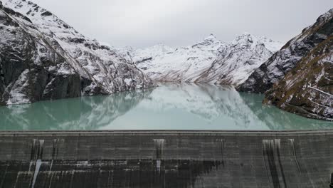 Erstaunliche-Drohnenaufnahmen-über-Einem-Alpensee,-Einem-Staudamm-Und-Einem-Riesigen-Alpenreservoir,-Gesäumt-Von-Schroffen-Bergen,-Mit-Einem-Uferpfad-Nach-Dem-Ersten-Schneefall-Im-Winter-2022