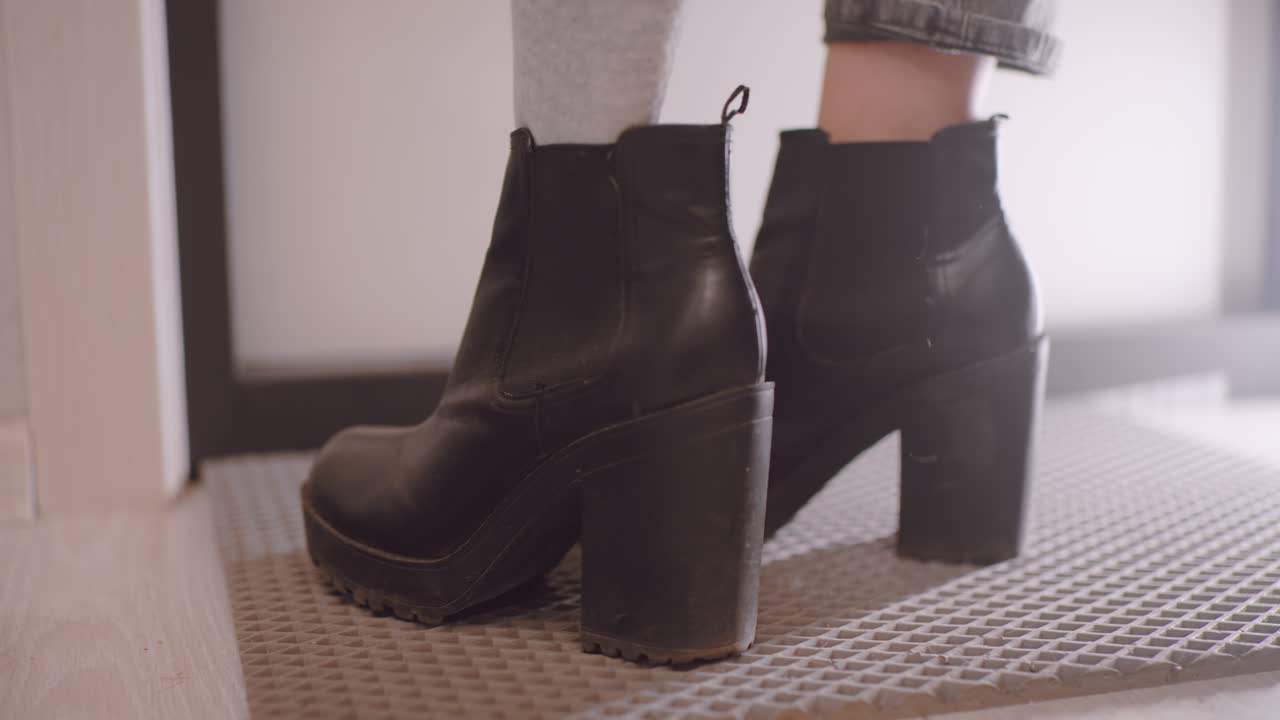 Close up of female in socks stepping into black leather boots with high chunky heels, preparing to leave home, showing detail of footwear and simple everyday action of putting on shoes
