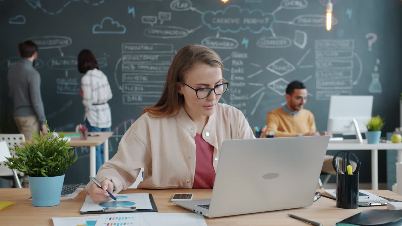 mujer joven trabajando con una computadora portátil en la oficina mientras sus colegas escriben en la pizarra en el fondo