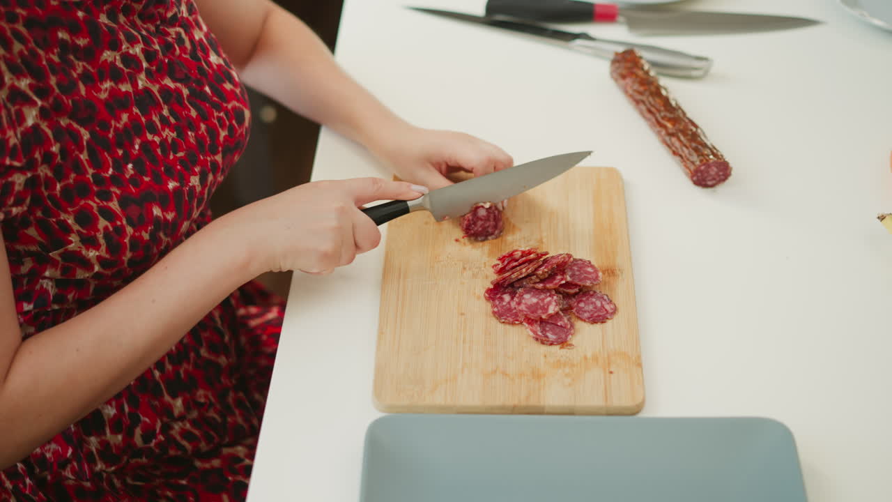 Confectioner hands skillfully slicing vibrant red cured meat into uniform thin rounds on wooden board over bright white table, emphasizing precise technique, glossy marbled texture, kitchen setting