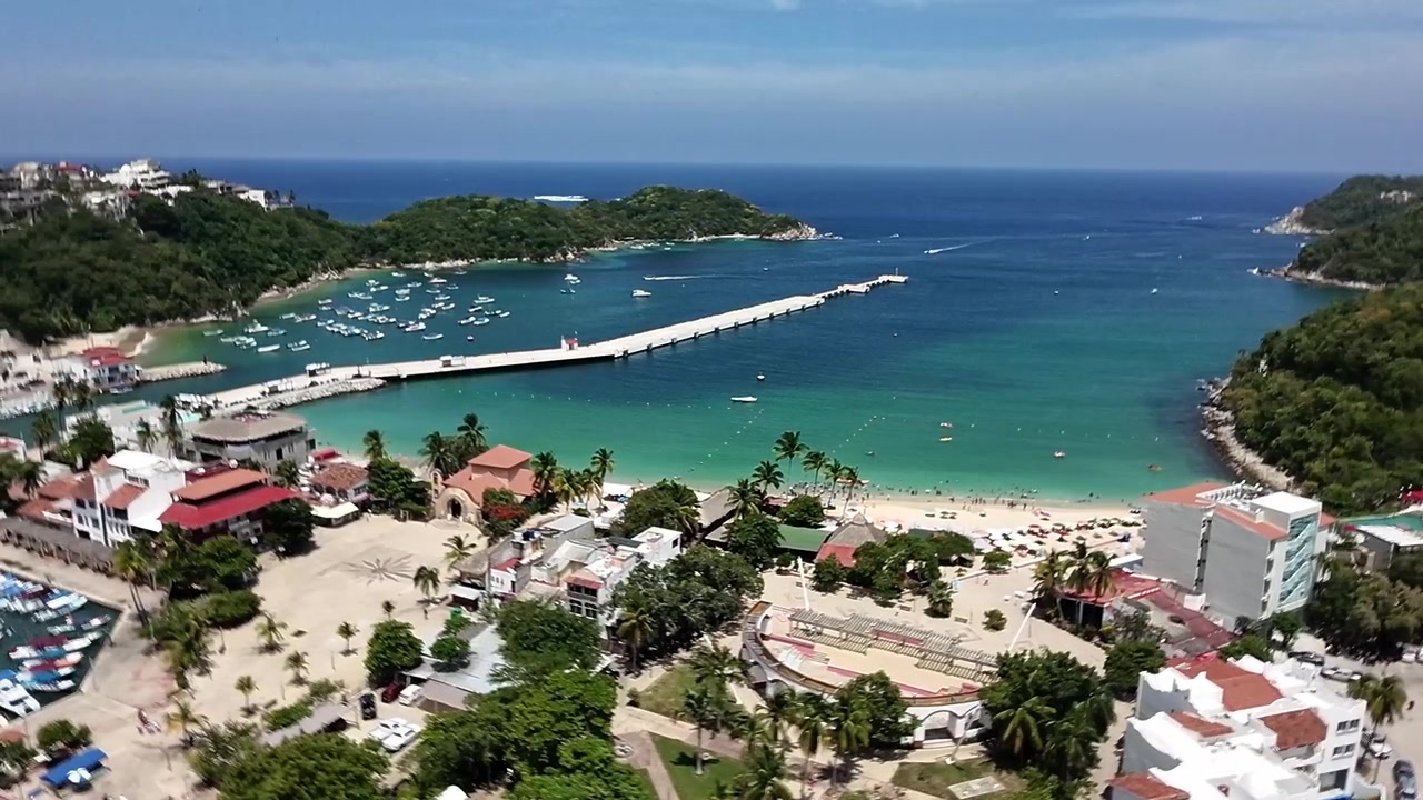 Ripresa aerea con drone di una baia tropicale con acqua turchese, spiaggia sabbiosa, marina, barche, palme ed edifici costieri a Huatulco, Messico, sotto un cielo azzurro e limpido