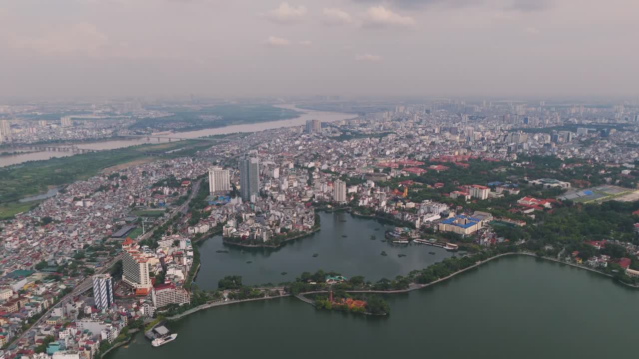 Wide drone shot of Hanoi's West Lake (Tay Ho), showing the West Lake and the surrounding cityscape with high-rise buildings and a mix of urban development. Vietnam UHD