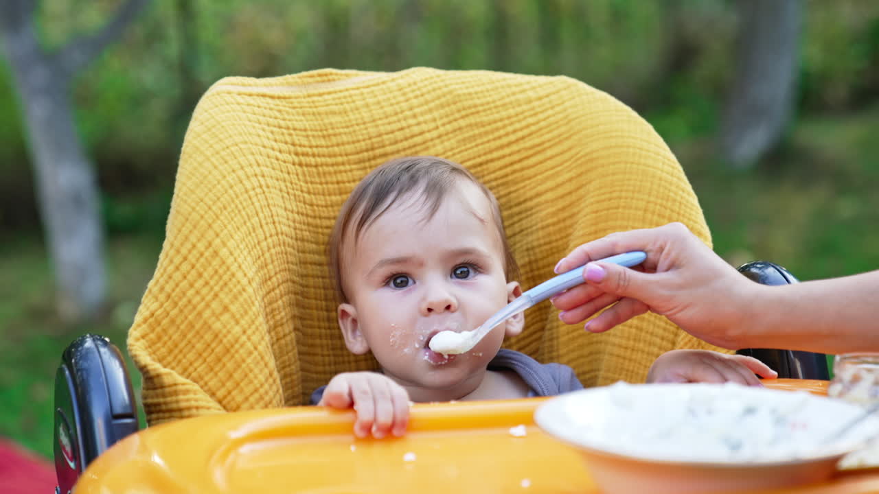 Loving mom giving food from spoon to a baby in yellow chair. Baby nutrition outdoors in the garden. Blurred nature backdrop.