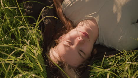 young woman lying peacefully in grassy field, surrounded by tall green blades of grass, with sunlight casting gentle shadows on her face as she blinks her eyes gently