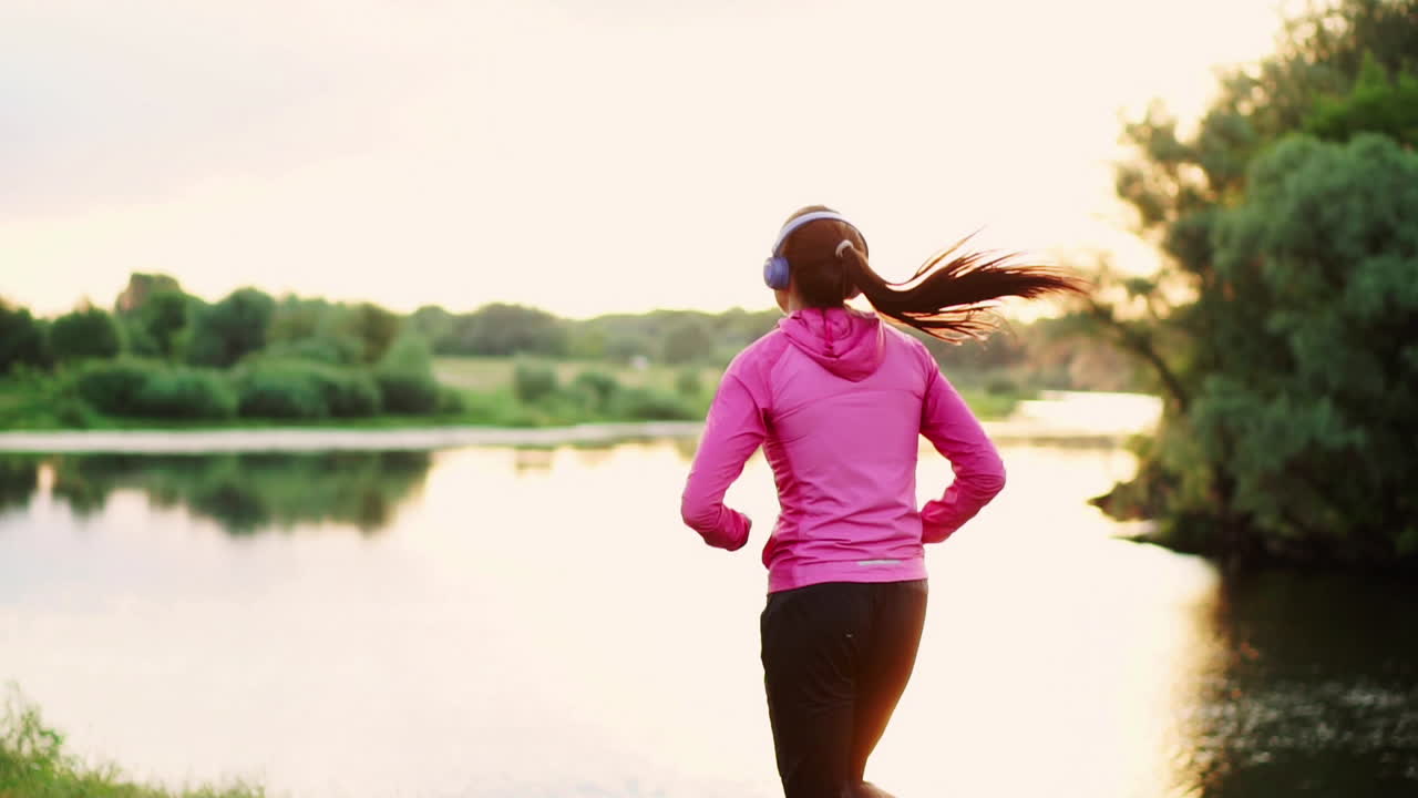 Brunette with long hair in headphones runs along the river in the Park in the morning at sunrise in the summer in a pink jacket and black pants