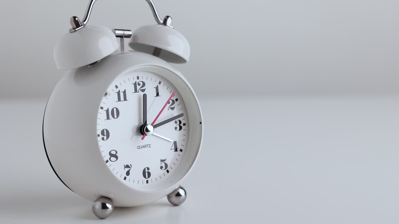 Time lapse movie. Small white alarm clock, black numbers, set the time for 12.00 o'clock, placed on a white table.