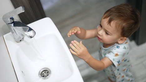 adorable toddler putting the hands under water tap. lovely kid playing with water in bathroom. top view.