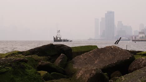 View-of-Manhattan-covered-in-smoke-from-wildfires-seen-from-beach-on-the-east-river-with-mossy-rocks-in-the-foreground