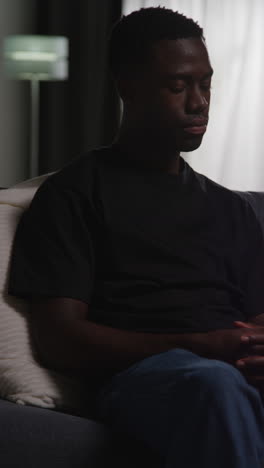 vertical video of unhappy and depressed young man sitting on sofa at home looking anxious and worried resting head on hand 2