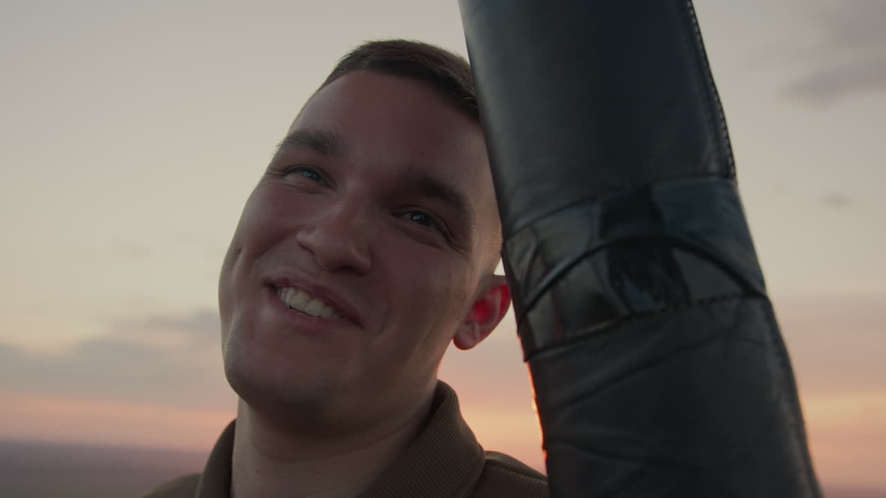 young man interacts with camera from hot air balloon basket as warm sunset light bathes smiling face against pastel sky over sprawling field, conveying sense of adventure freedom