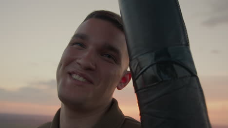young man interacts with camera from hot air balloon basket as warm sunset light bathes smiling face against pastel sky over sprawling field, conveying sense of adventure freedom