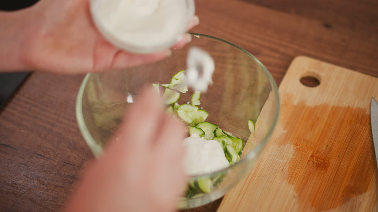 close up of person holding small bowl and spoon while adding creamy white dressing into sliced cucumber placed in transparent glass bowl beside cutting board with kitchen knife on wooden surface