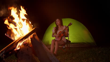 asiatic woman musician playing guitar ukulele in front at bonfire while camping outdoor at night