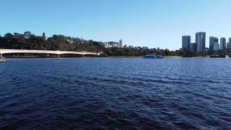 Drone-Flying-across-Swan-River-over-cruise-boat-to-Narrows-Bridge,-Freeway-and-Kings-Park-with-Perth-Skyline