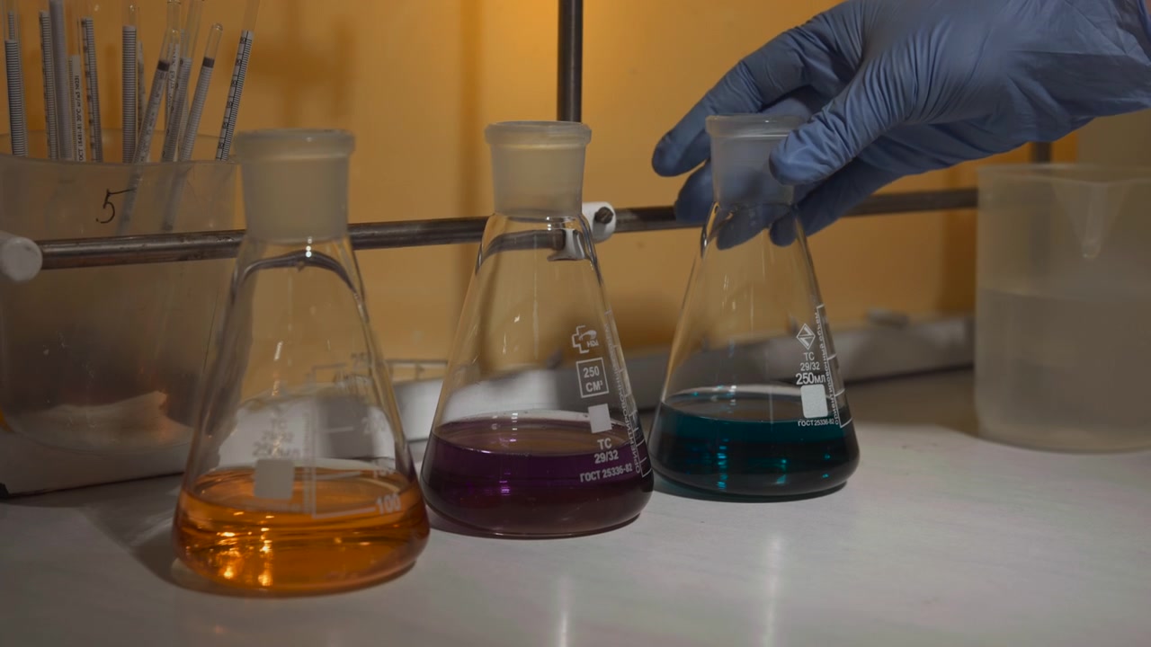 Mano guantata si allunga per afferrare e sollevare la beuta di Erlenmeyer più a destra contenente una soluzione blu, mentre due beute vicine con liquidi arancioni e viola rimangono su un bancone bianco in un ambiente di laboratorio luminoso