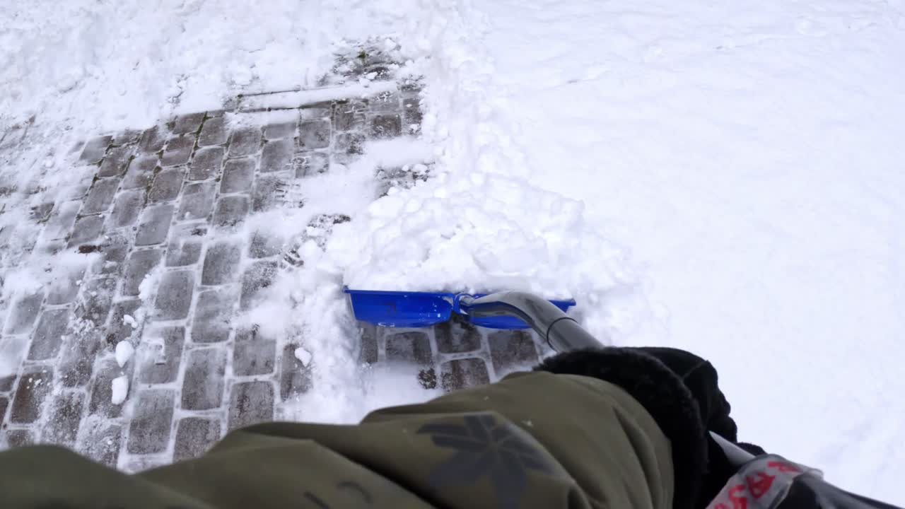 POV (synspunkt) skubber en blå skovl langs kanten af en brostensbelagt indkørsel, der skærer en skarp kant gennem dyb sne og efterlader et rent spor ved siden af et snedækket felt med fodspor.