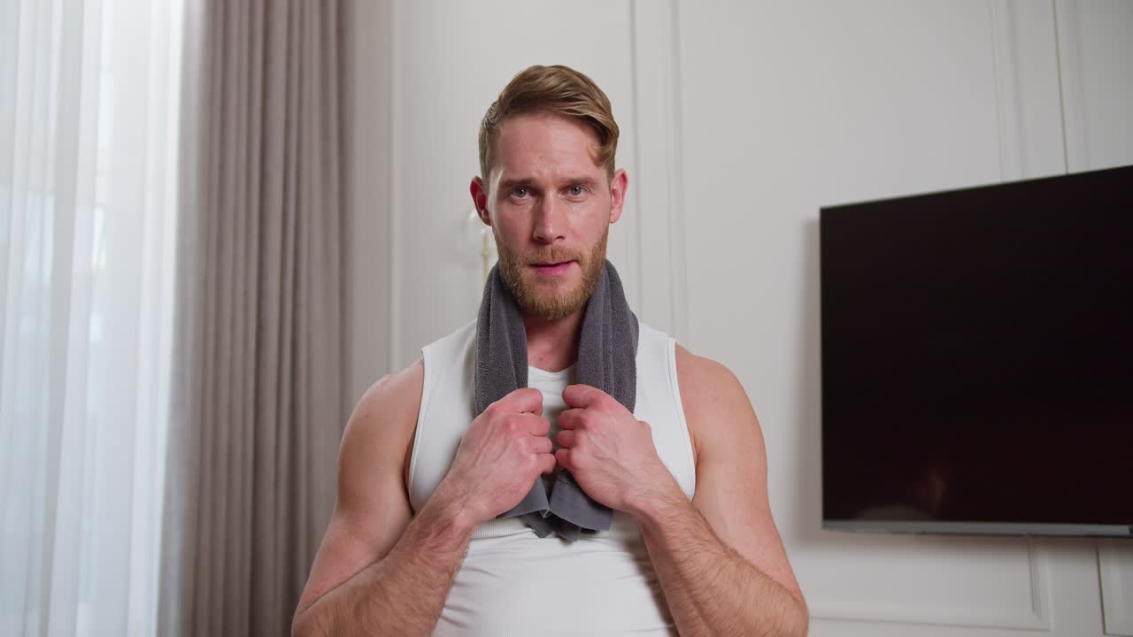 색 티셔츠를 입은 갈색 머리카락과 운동 체격을 가진 행복하고 자신감있는 남자의 초상화, 현대적인 아파트에서 워밍업하는 동안 목에 회색 수건을 들고 집에서 운동합니다.