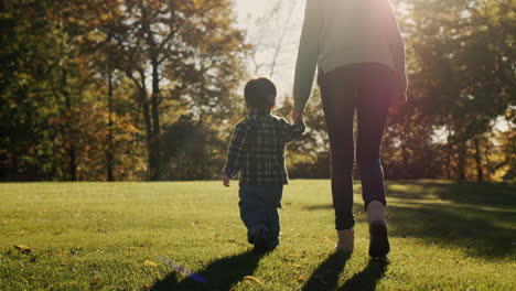 Mom-leads-the-baby-by-the-hand,-walking-next-to-the-green-grass-in-the-rays-of-the-setting-sun.-Happy-motherhood-and-childhood.