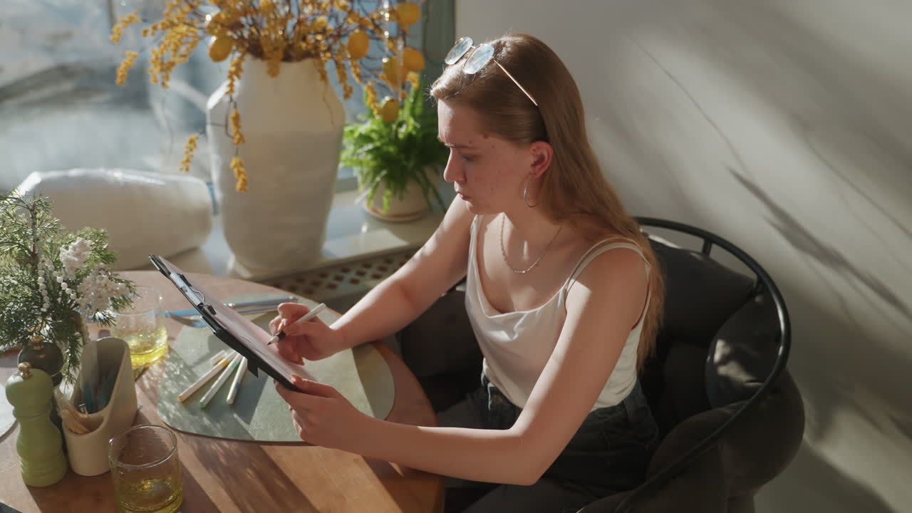 Young white woman using tablet at table, freelancer planning creative menu, stylus in hand, sunlit dining nook with vase and plants, casual summer outfit and sunglasses on head, focused planning flow