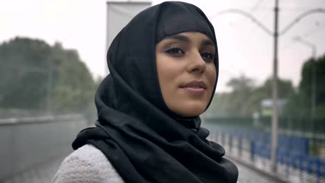 young muslim woman in hijab is waiting for train on railway station, raining, religion concept, urban concept. weather concept, side view