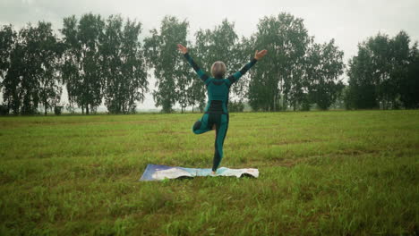vista trasera de una mujer de pie con la pierna derecha en una alfombra de yoga colorida en postura de árbol, practicando yoga en un vasto campo de hierba con árboles en el horizonte, manteniendo el equilibrio y la calma bajo un cielo nublado