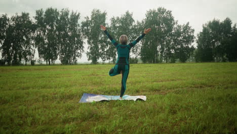 back view of woman standing with her right leg on colorful yoga mat in tree pose, practicing yoga in a vast grassy field with trees on the horizon, maintaining balance and calm under a cloudy sky