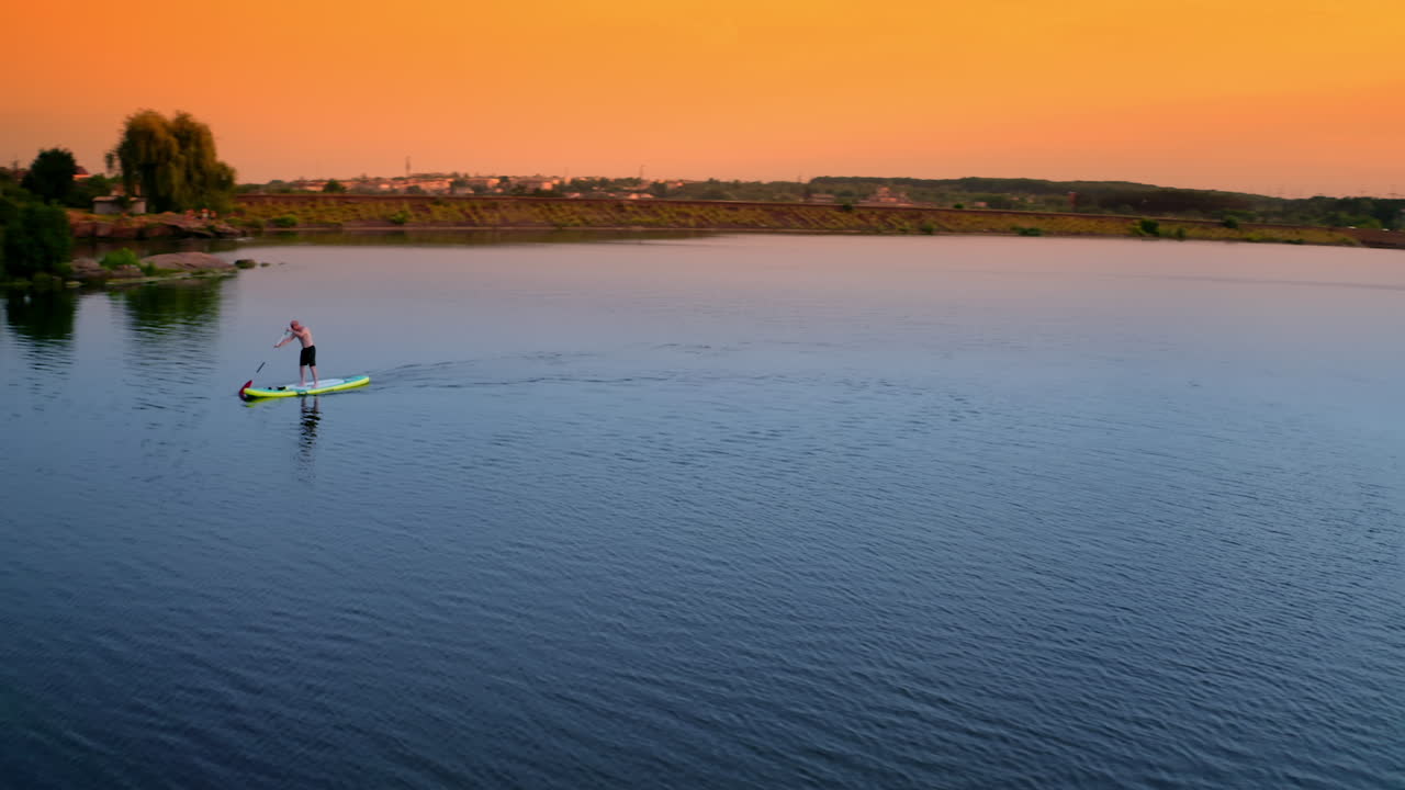 Sportsman on a paddle boat on lake at sunset. Man sailing on a board with oar along the evening river in the countryside. Extreme sport on water.