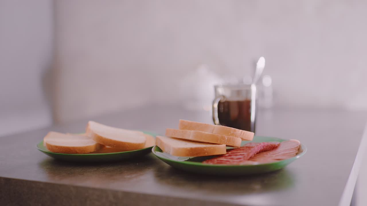 Hand carefully places green plate with slices of bread and meat on table beside steaming coffee cup, capturing domestic breakfast preparation in warm indoor atmosphere