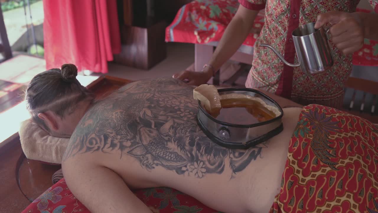 Spa therapist pours warm oil into the wax-lined container on the back of a male patient enjoying a Kati Vasti Ayurvedic treatment