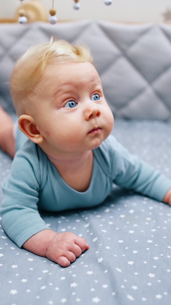 Liten babygutt med store blå øyne ligger på magen. Søt spedbarn i blå body i barnesengen. Vertikal video