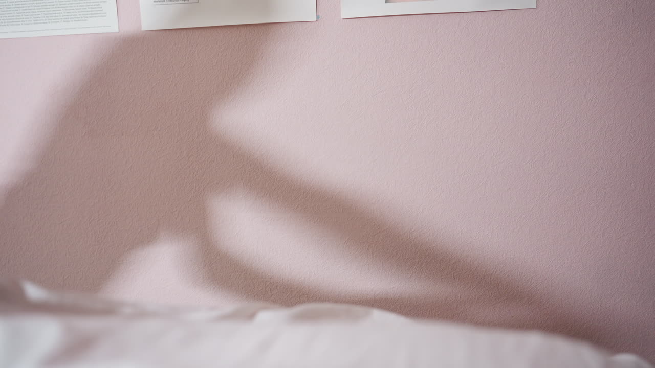 Shadow of massage therapist extending arms during massage session, softly cast on pink wall near anatomical sketches, with part of client body under white sheet visible in lower frame