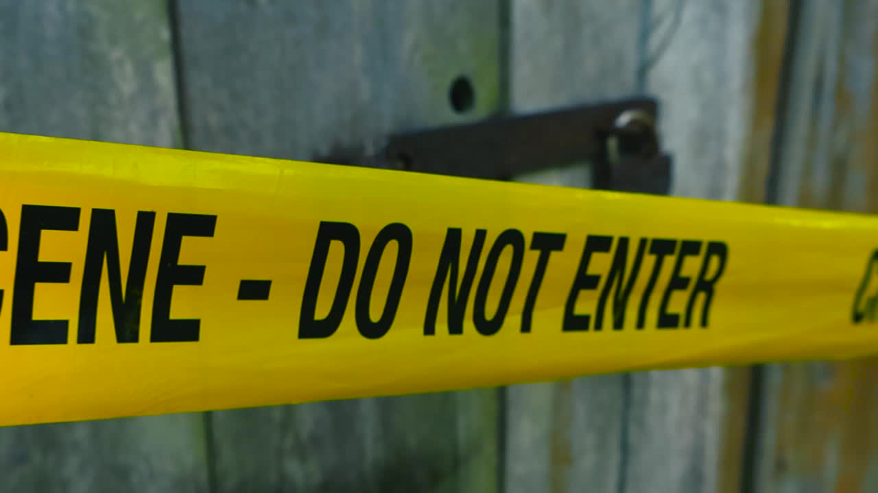 Close up view of a yellow colored police forensic investigation Crime Scene tape line ribbon in front of a wooden barn or farm house door with a rusty padlock in front of it locked. Countryside, day