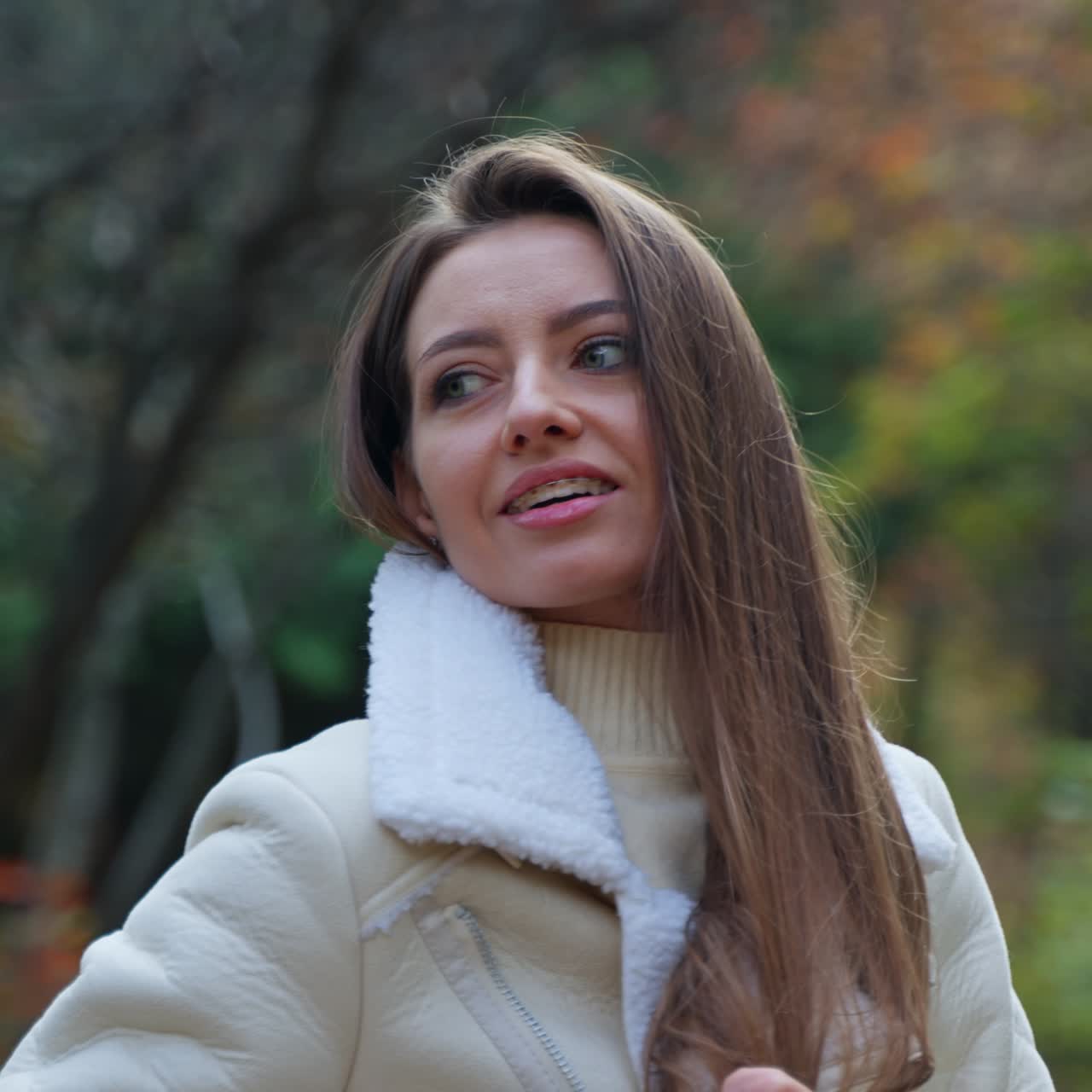Brunette is cold a little and tries to warm up coating in her jacket. Smiling lady looks aside standing in the autumn forest