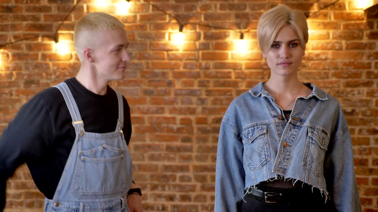 joven hermosa chica tranquila está parando y extraordinario chico hipster está bailando a su lado, mirando a ella, fondo de ladrillo