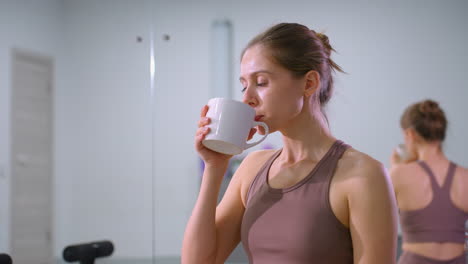 lady takes break from exercise, sipping coffee while seated in gym, large mirror behind reflects her relaxed posture, gym equipment and fitness mats add to wellness atmosphere