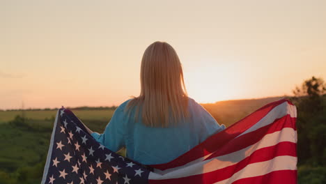 Freihandaufnahme:-Eine-Frau,-Die-Mit-Einer-Amerikanischen-Flagge-Auf-Den-Schultern-Läuft,-Sieht-Zu,-Wie-Die-Sonne-über-Einem-Weizenfeld-Untergeht.