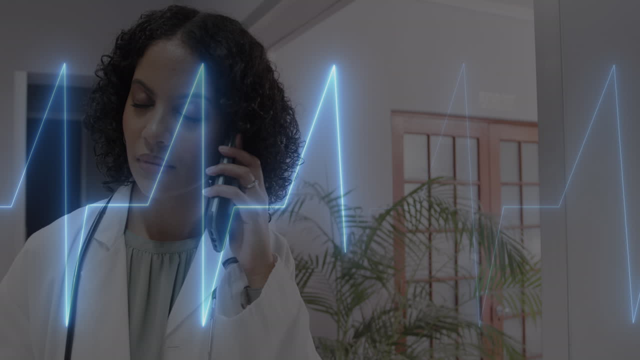 Animation of cardiograph over happy biracial female doctor talking on smartphone