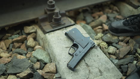 a close view of a handgun placed on the concrete base of a railway track surrounded by rocks, with view of someone leg