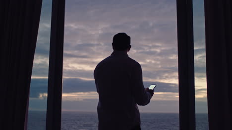 young-man-using-smartphone-in-hotel-room-texting-sharing-vacation-lifestyle-on-social-media-enjoying-view-of-ocean-at-sunset