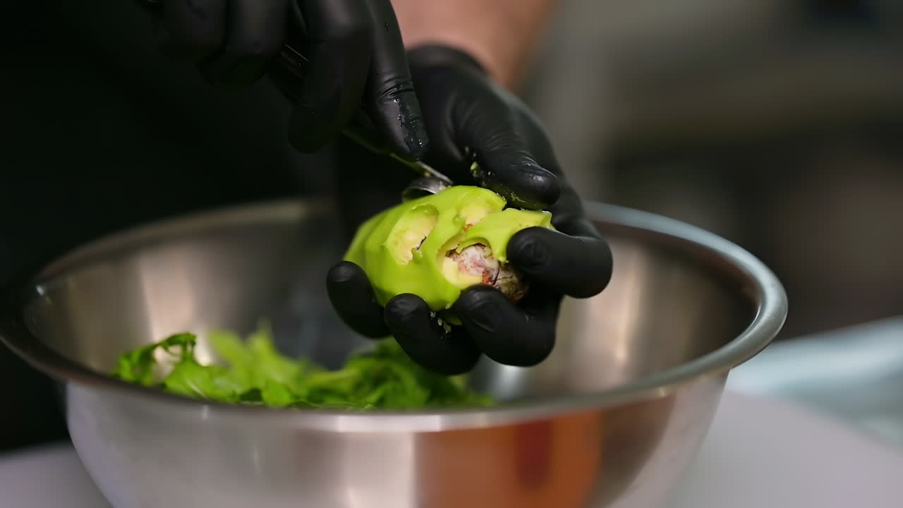 Caucasian man wearing black gloves preparing food in restaurant kitchen. Male putting avocado at the salad with special spoon. Cooking vegetarian food at modern restaurant kitchen