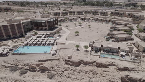 Drone-flies-over-a-beautiful-architecture-stone-built-beresheet-hotel-in-a-desert-landscape,-Flying-back-and-reveals-the-beautiful-cliff