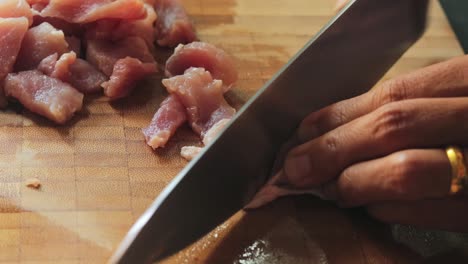 carefully slicing raw pork meat into small pieces on a bamboo chopping board with a steel knife - close up, side view